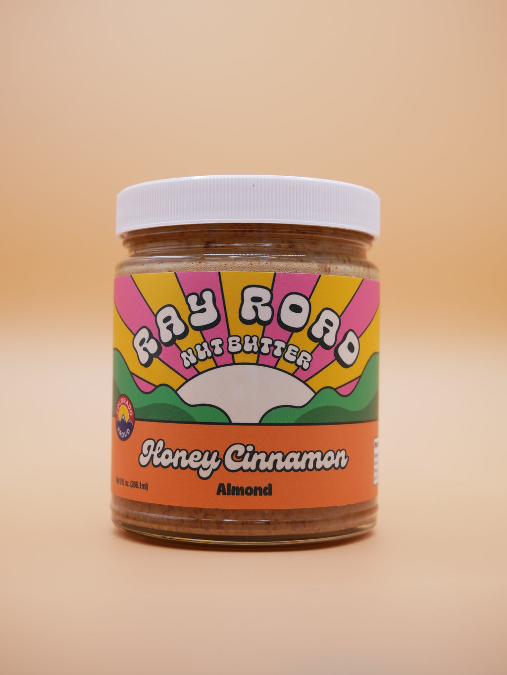 A jar of Fay Road Nut Butter Honey Cinnamon Almond on a solid beige background. The jar has a white lid and a colorful retro-style label with pink, yellow, green, and orange sections.