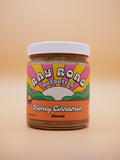 A jar of Fay Road Nut Butter Honey Cinnamon Almond on a solid beige background. The jar has a white lid and a colorful retro-style label with pink, yellow, green, and orange sections.