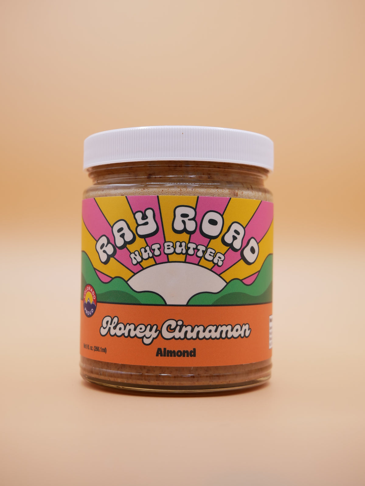 A jar of Fay Road Nut Butter Honey Cinnamon Almond on a solid beige background. The jar has a white lid and a colorful retro-style label with pink, yellow, green, and orange sections.
