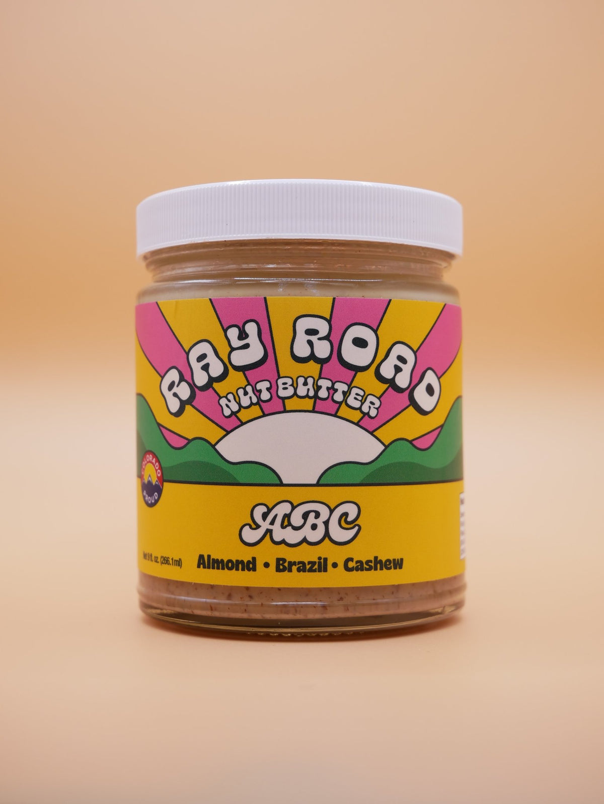 A jar of ABC Ray Road Nut Butter (Almond, Brazil, Cashew) on a solid beige background. The jar has a white lid and a colorful retro-style label with pink, yellow, green, and white graphics.