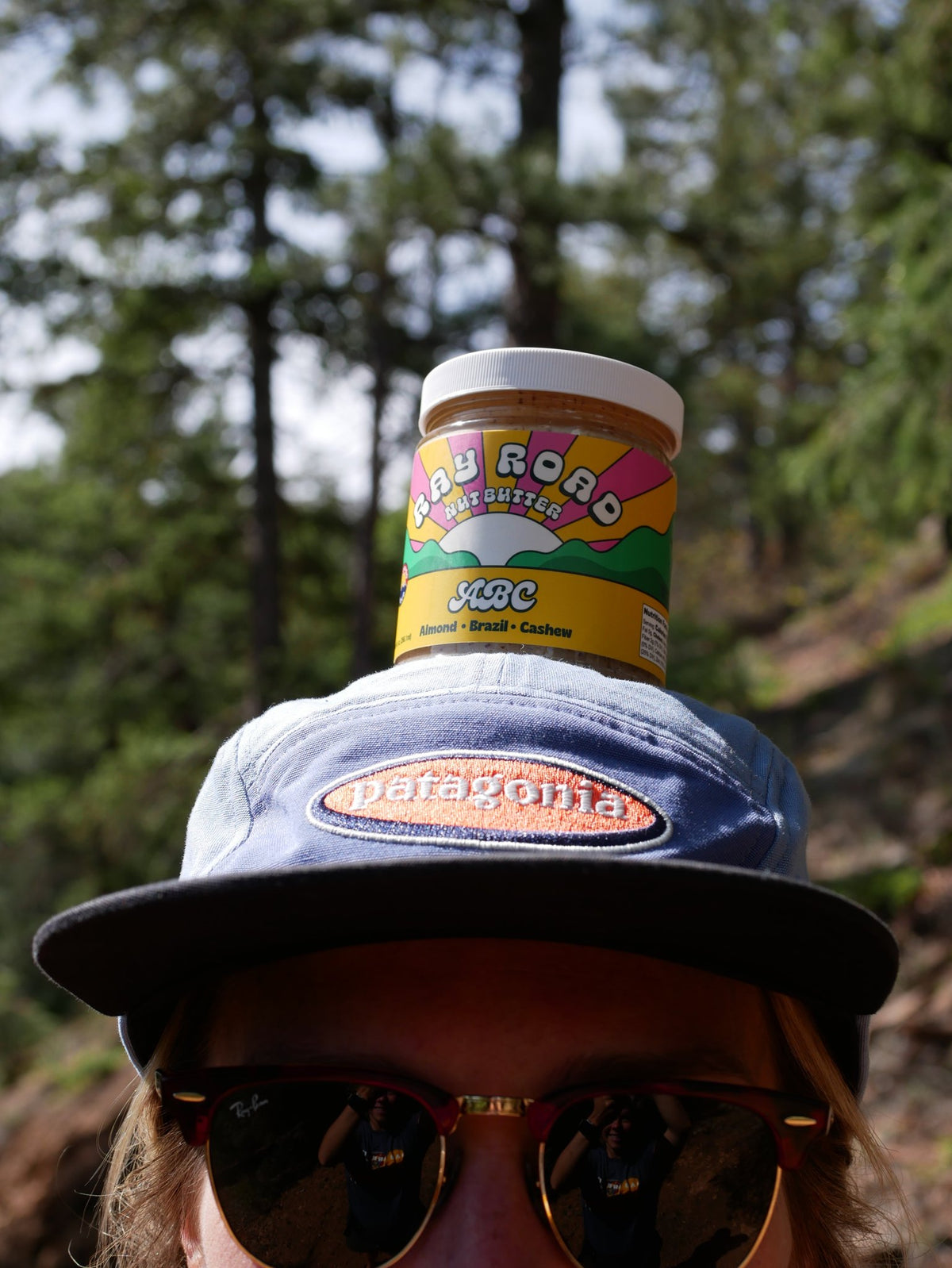 A person outdoors wearing a blue and black Patagonia cap with a jar of Robo Roar Spread balanced on top of their head. The background is a blurred forest.