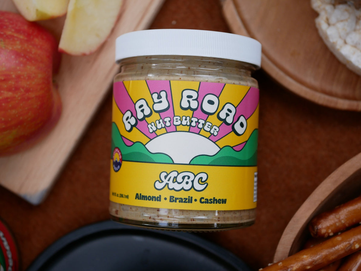 A jar of Ray Road Nut Butter (ABC Almond + Brazil + Cashew) centered in a styled flat-lay with snacks like sliced apples, pretzels, and crackers on wooden boards.