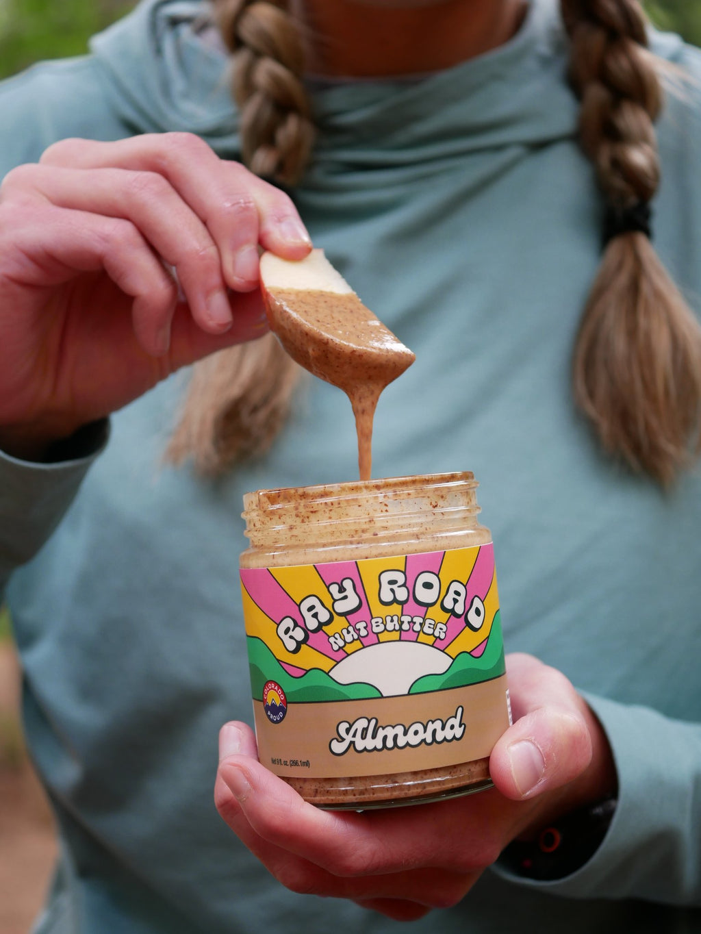 jar of ray road nut butter almond butter held in someones hand. the other hand holds an apple slice above the jar with nut butter dripping off. 