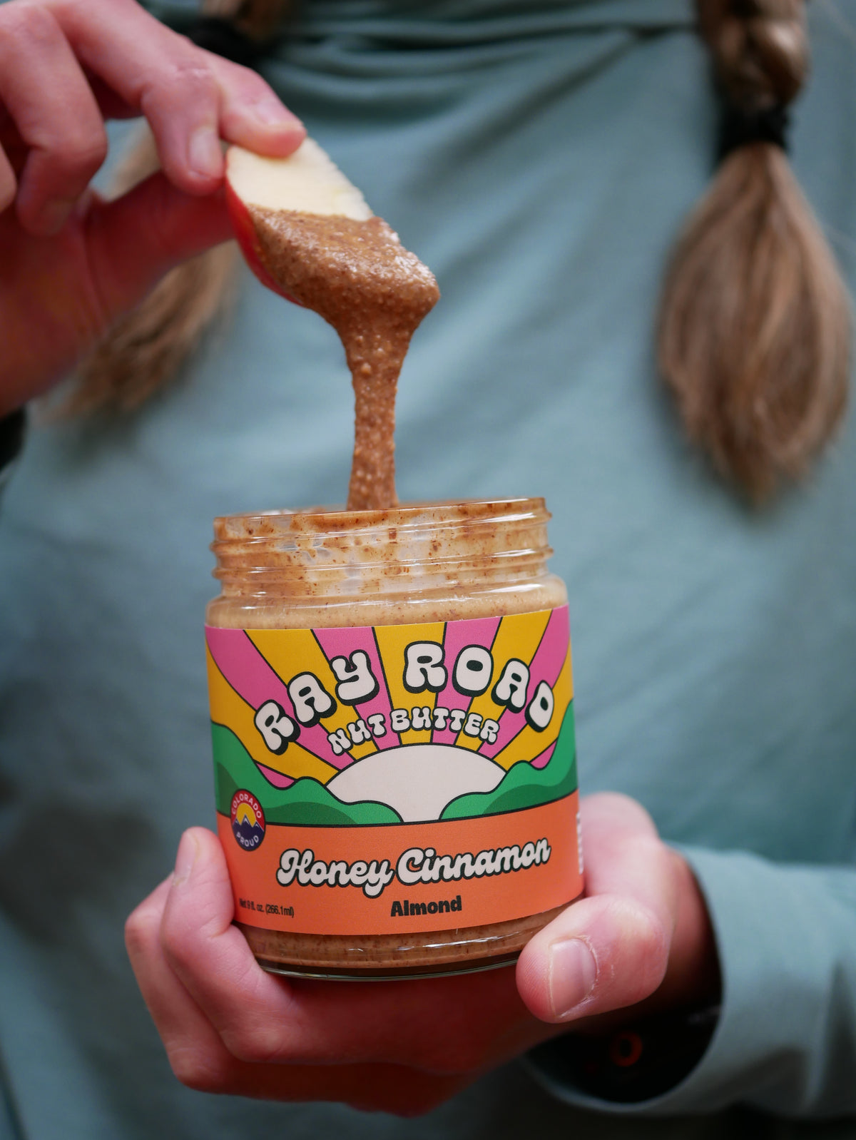 A person holding a jar of Ray Road Nut Butter Honey Cinnamon Almond butter while dipping an apple into the spread. The jar features a colorful retro label with pink, yellow, green, and orange sections.