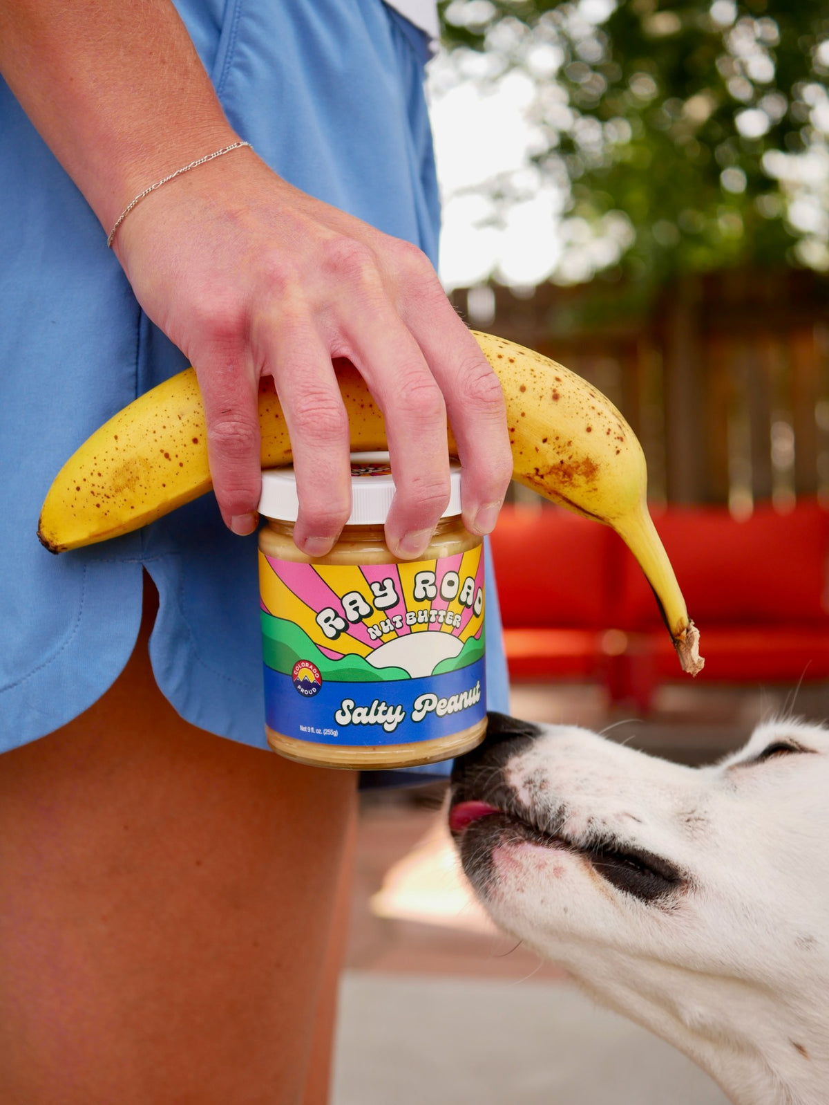 Hand holding a Salty Peanut jar and a banana while a dog sniffs the jar