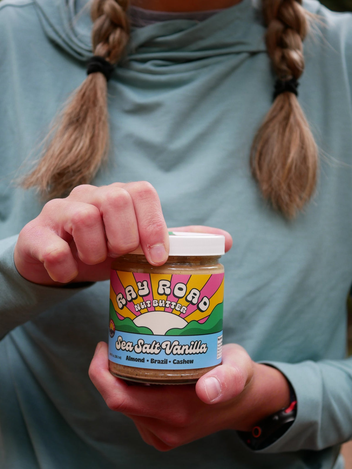 a person holding a jar of ray road sea salt vanilla nut butter, held in two hands, mid opening the jar