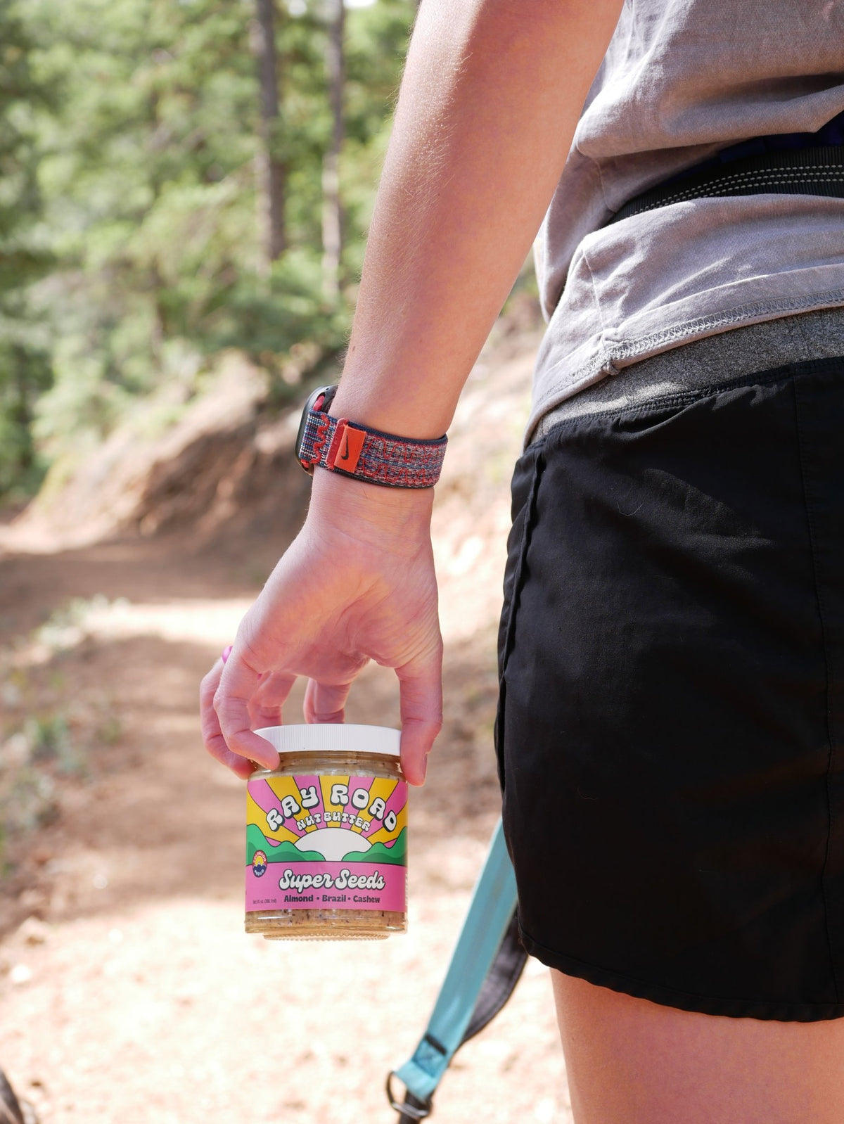 A person outdoors on a trail holding a small jar of 'Any Goes Gourmet' Jujube Nectar. The person is wearing a gray t-shirt, black shorts, and a patterned orange and black wristband.