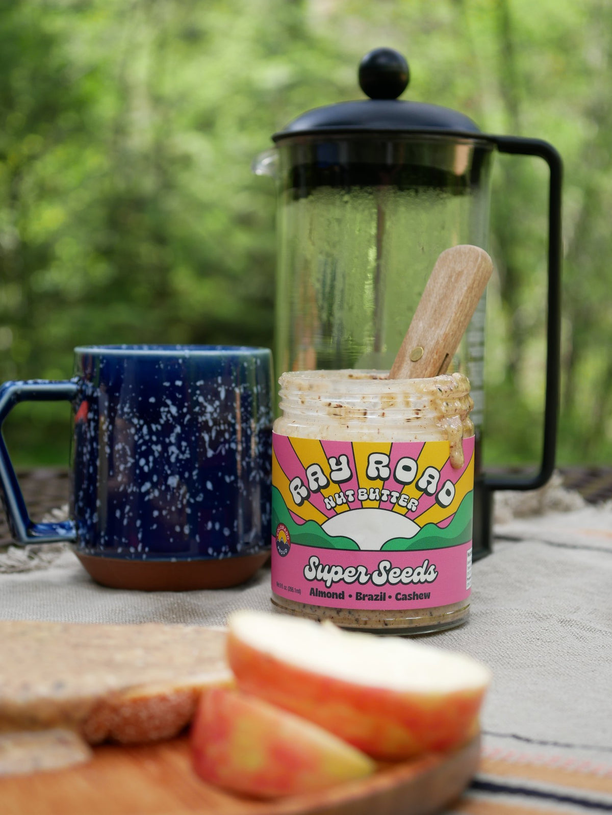 A jar of Ray Road Butterfused Super Seeds almond butter sits on a picnic blanket outdoors, surrounded by a blue speckled mug, a glass French press, and slices of bread and apple in a blurred green forest setting.