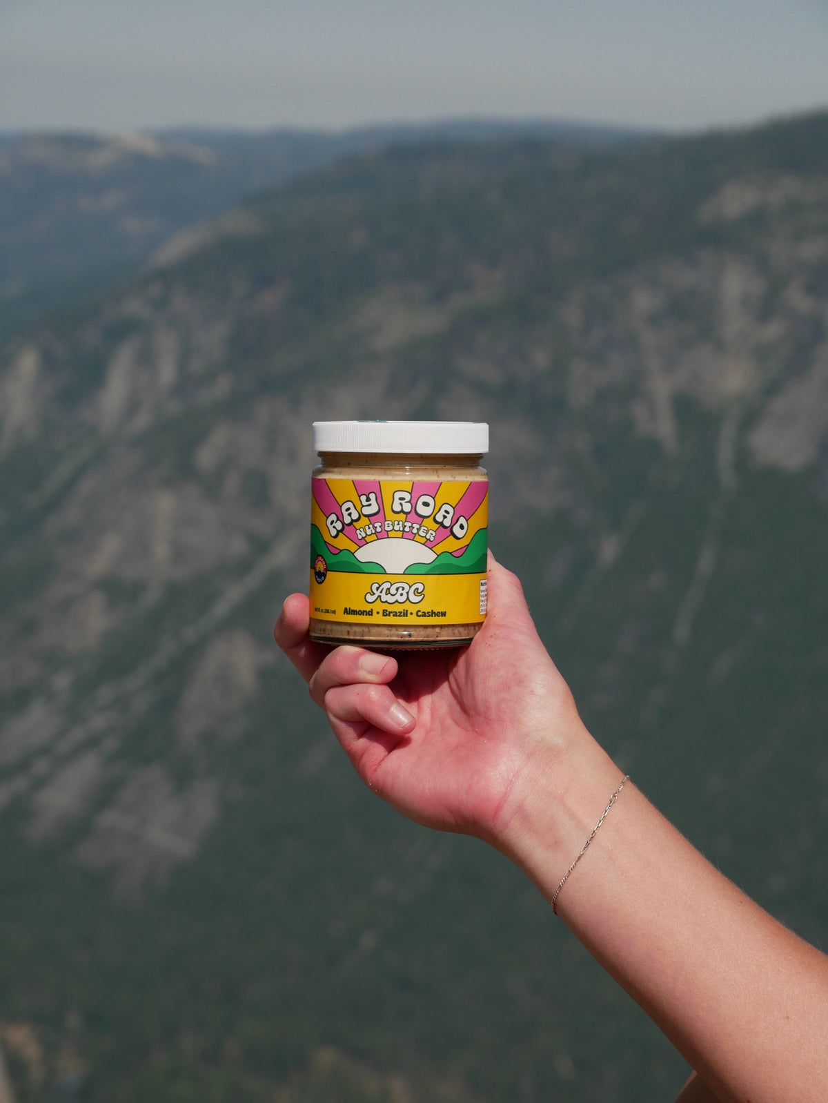 A hand holds up a jar of Ray Road nut butter above a hazy mountain valley